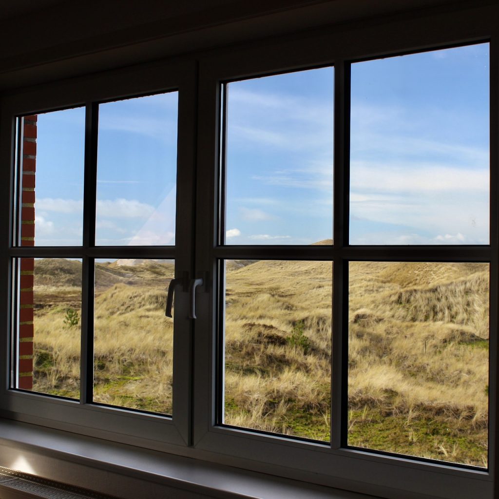 Blick aus einem weißen Sprossenfenster der AOK-Nordseeklinik auf die Dünenlandschaft. Darüber blauer Himmel mit leichten Wolkenschleiern.