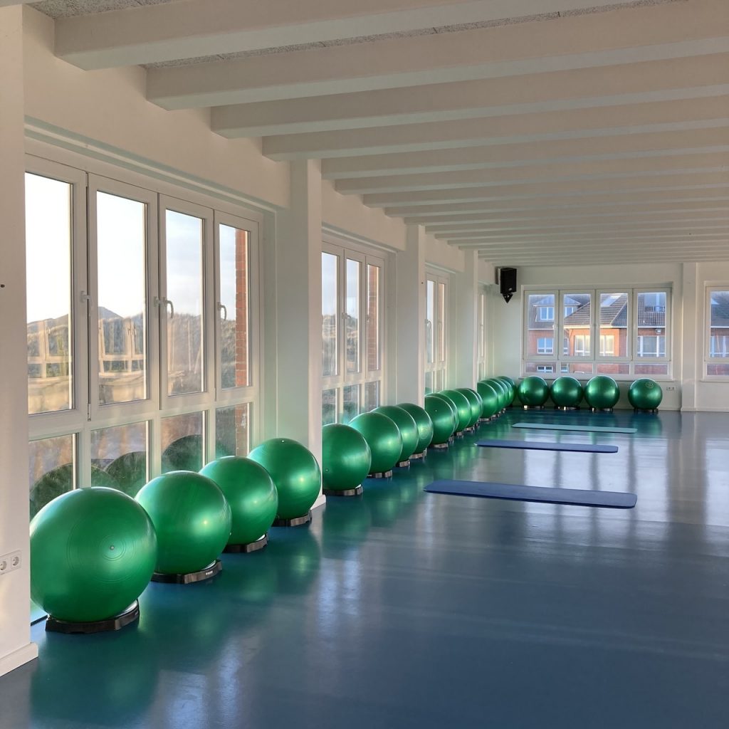 Die kleine Sporthalle der AOK-Nordseeklinik. An der großen Fensterfront sind grüne Bälle aufgereiht, die sich auf dem blauen Boden spiegeln. Durch die Fenster sieht man die Dünen und das Dünenhaus.