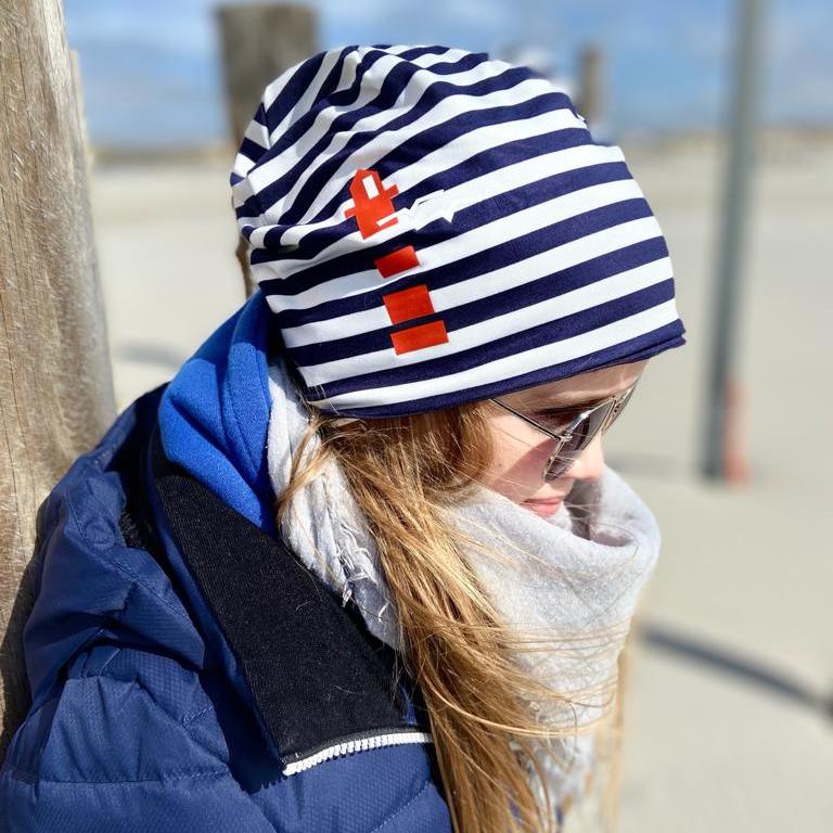 Ein Kind lehnt am Strand an einem Holzpfahl. Es trägt eine blau-weiß-gestreifte Beanie auf dem Kopf.