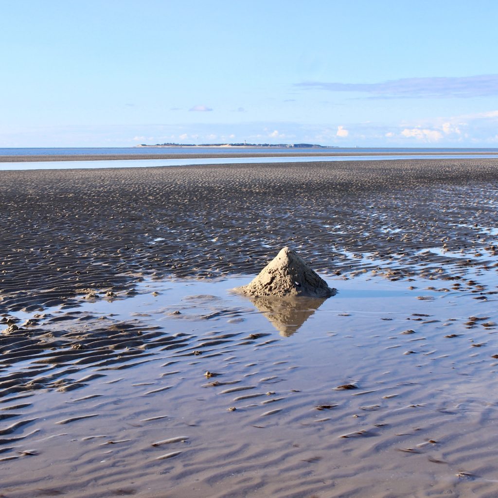 Das Watt der Nordsee bei Ebbe. Im Vordergrund eine kleine Sandpyramide, die sich im Wasser spiegelt. Am Horizont die Insel Sylt.