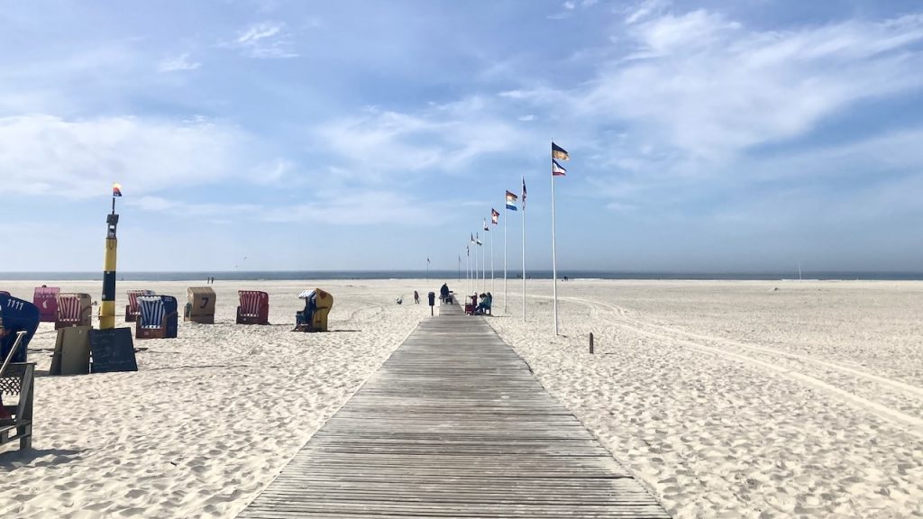 Breiter Strand von Norddorf, in der Mitte ein Bohlenweg, gesäumt von Flaggen, links einige Strandkörbe